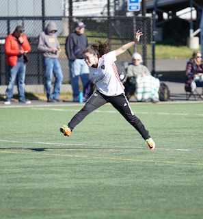 A person is mid-air on a sports field, appearing to be involved in an athletic activity. Several spectators are on the sidelines, some seated and others standing, with one individual capturing the event with a camera. The background includes a fence and various casual observers, some of whom are wearing winter clothing.