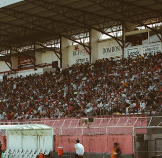 A crowded stadium with spectators filling the stands, observing an event. The stadium is covered and has multiple sections labeled with various advertisements visible on the walls. In front of the stands, a few people are gathered near an area with chairs and a white canopy.